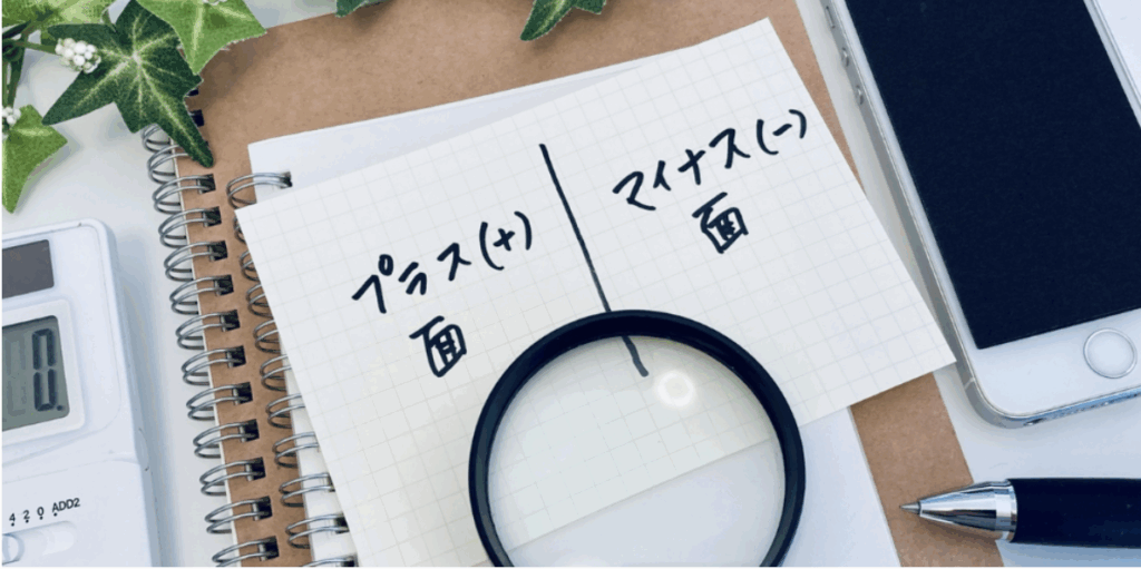 方眼紙に「プラス（＋）面」「マイナス（−）面」と手書きし、中央にルーペを置いて長所・短所を比較検討するワークシート。周囲に電卓・ノート・スマホ・ペンと観葉植物