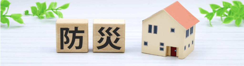 木製ブロックに黒文字で「防災」と表記し、隣にミニチュア住宅を配置。背景のグリーンが家庭の防災対策を連想させるビジュアル