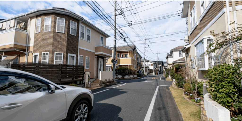 閑静な住宅街の曲がり角を白いSUVが走り抜ける様子。両側の洋風戸建てと青空に伸びる電線が映える街並みイメージ