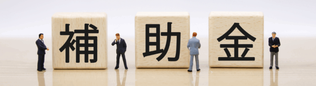 木製ブロックに黒文字で「補」「助」「金」と並び、その周囲にスーツ姿のミニチュアビジネスマンが立つ補助金イメージ