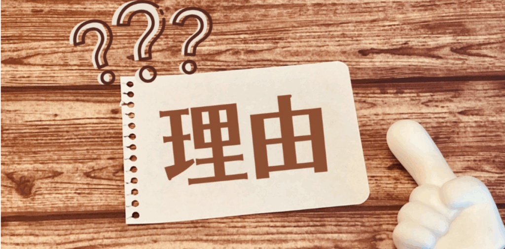 木目のテーブルに置かれたメモ用紙に「理由」と書かれ、クエスチョンマークと指差しの手が強調